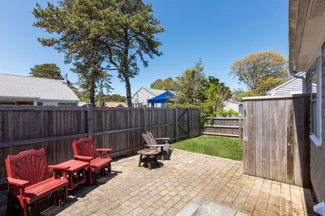 Brick paver patio with two red chairs, a small red table and a gray Adirondack chair, enclosed by a wooden fence with pine trees.