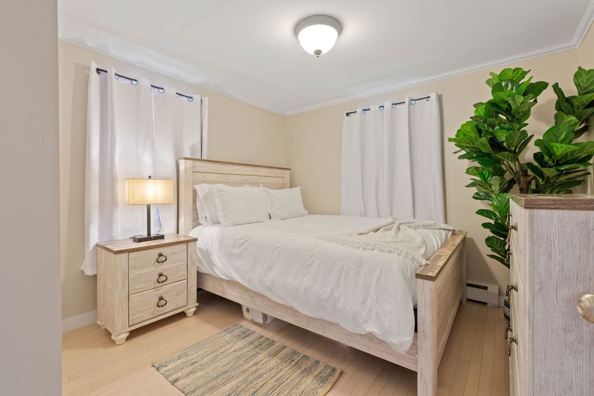 Bedroom with a queen bed in white linens, whitewashed nightstand and lamp, dresser, potted plant, two curtained windows and a rug.