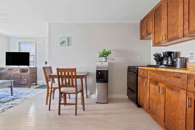 Open-plan room with wood kitchen cabinets, black range, coffee maker, water dispenser, a wood table with two chairs and a TV beyond.