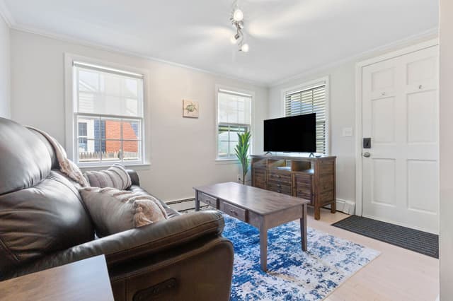 Living room with a leather reclining sofa, wood coffee table on a blue rug, TV on a wood console, three windows and a white door.