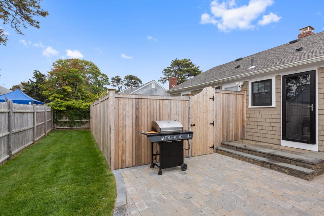 Paver patio with steps up to the back door, a gas grill by the fenced gate, and a strip of lawn along the fence.