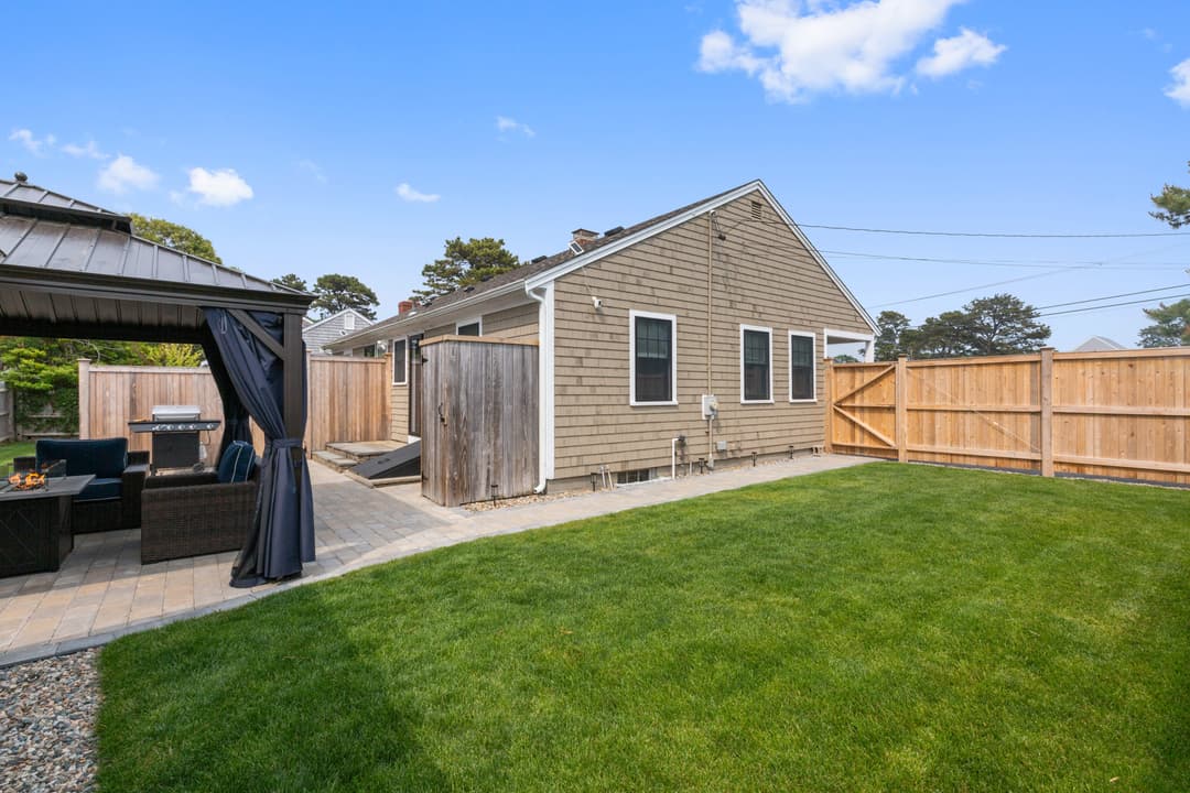 Fenced grass backyard beside a cedar-shingled single-story house, with a gazebo, grill and outdoor shower stall at left