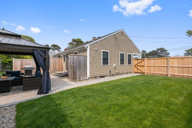 Fenced grass backyard beside a cedar-shingled single-story house, with a gazebo, grill and outdoor shower stall at left.