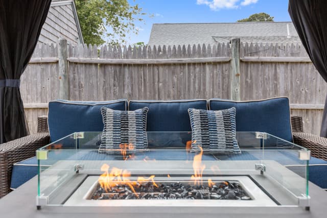 Close-up of a lit gas fire table with glass wind guard in front of a blue-cushioned wicker sofa with striped pillows.