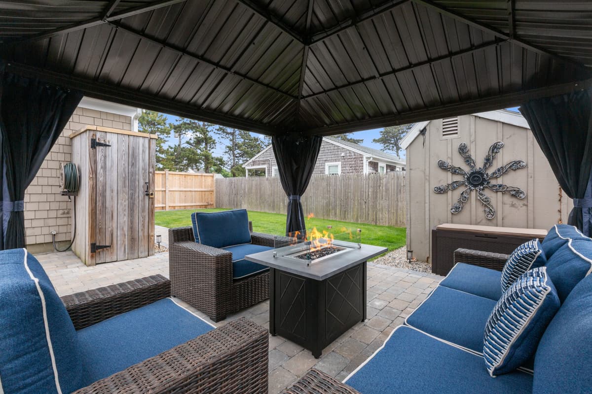 View from inside a metal-roof gazebo with blue-cushioned wicker sofa and chairs around a lit gas fire table, outdoor shower and shed beyond.
