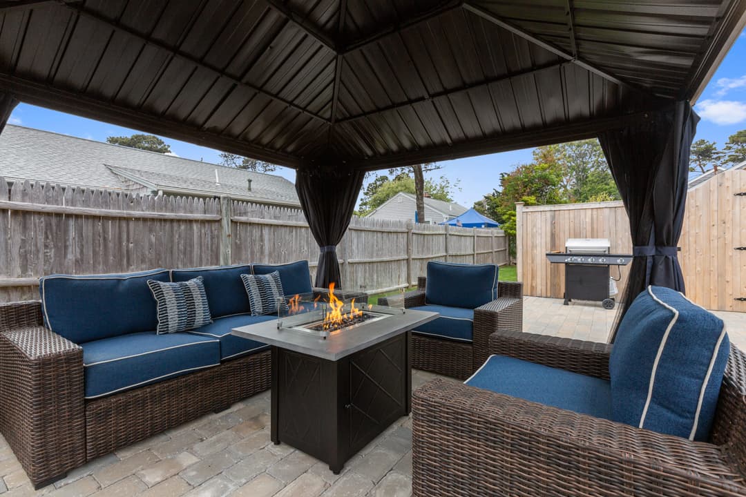 Covered gazebo on a paver patio with wicker sofa and chairs with blue cushions around a lit gas fire table; a grill sits by the fence.