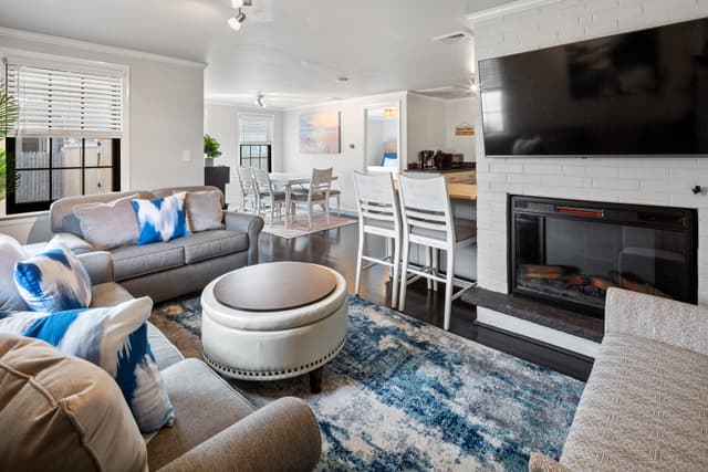 Living room with gray sofas, round ottoman, blue rug, and a wall-mounted TV over a white brick electric fireplace; dining area behind.