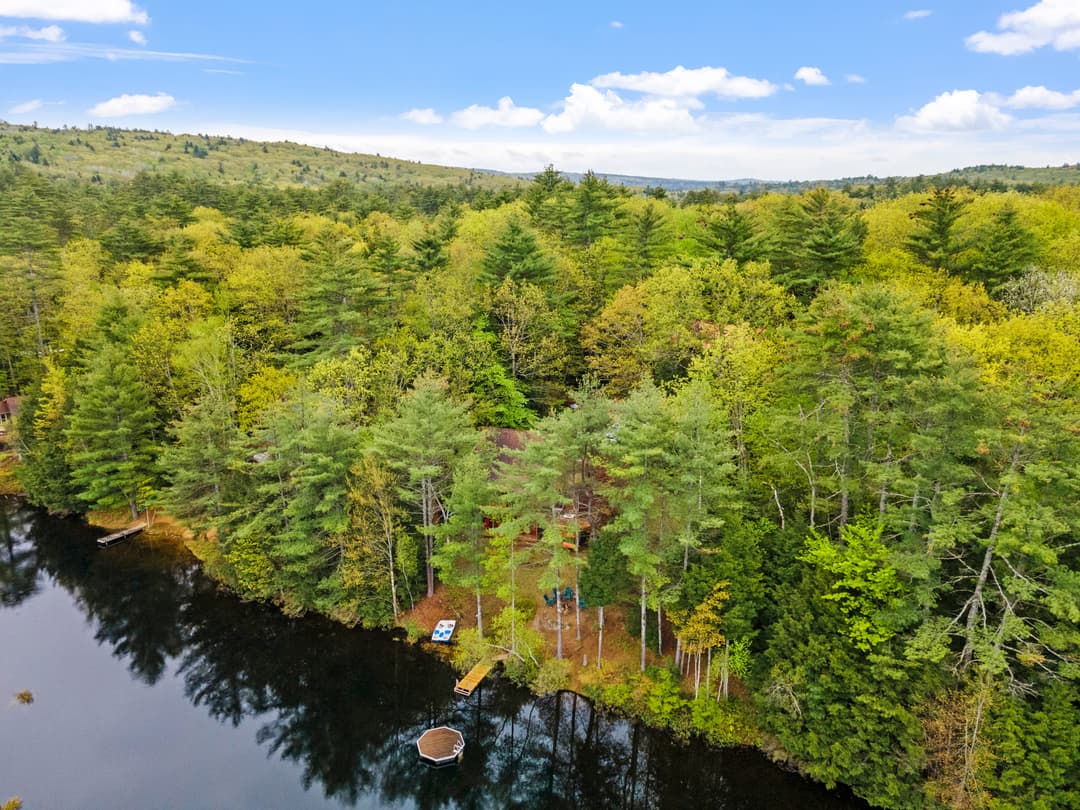 Aerial view of a wooded lakeshore with a small dock, a floating platform in the water and forested hills beyond.