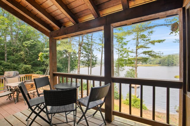 Covered porch with a wood ceiling, a small round table and three wicker chairs, overlooking a lake through posts.