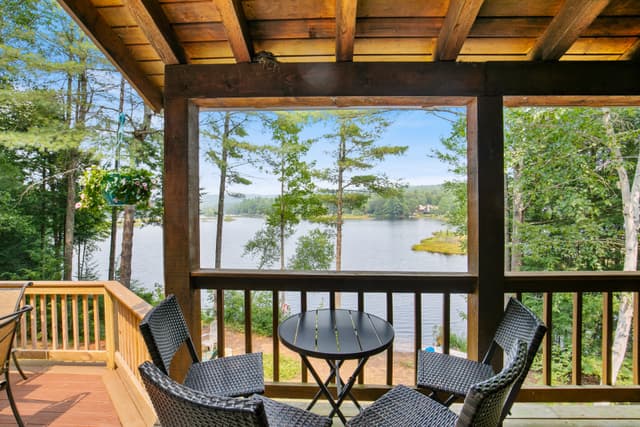 Round black table and four wicker chairs on a covered porch, with a lake framed between timber posts and a hanging plant.