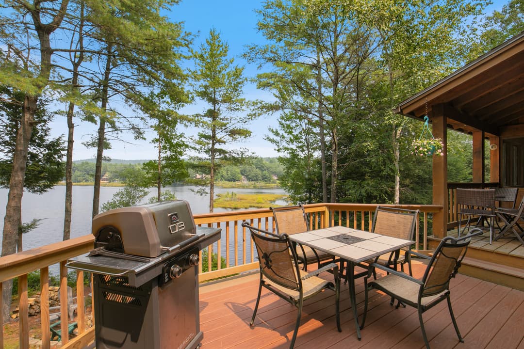 Open wooden deck with a gas grill and a tiled table with four chairs, trees and a lake beyond the railing.