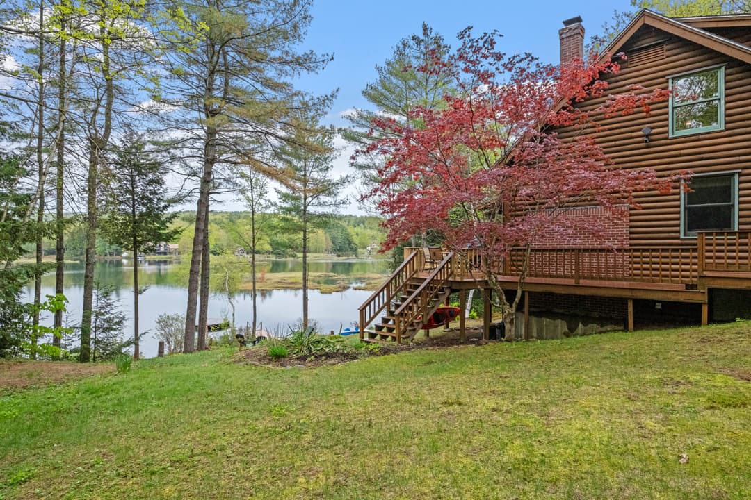 Side of a log home with a raised wooden deck and stairs, a red-leaf tree, and a lawn sloping down to the lake.