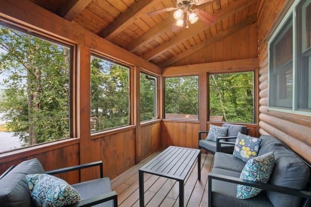 Screened porch with wood walls and ceiling fan, a gray sofa, two armchairs with patterned pillows and a black coffee table.