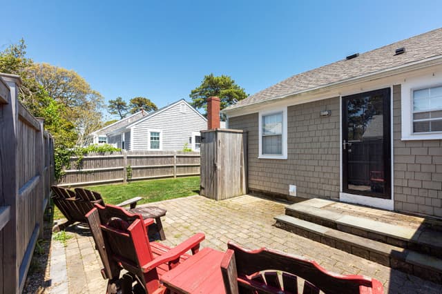 Paver patio with red and wooden Adirondack chairs beside a shingled cottage, a back door with steps and an outdoor shower stall.
