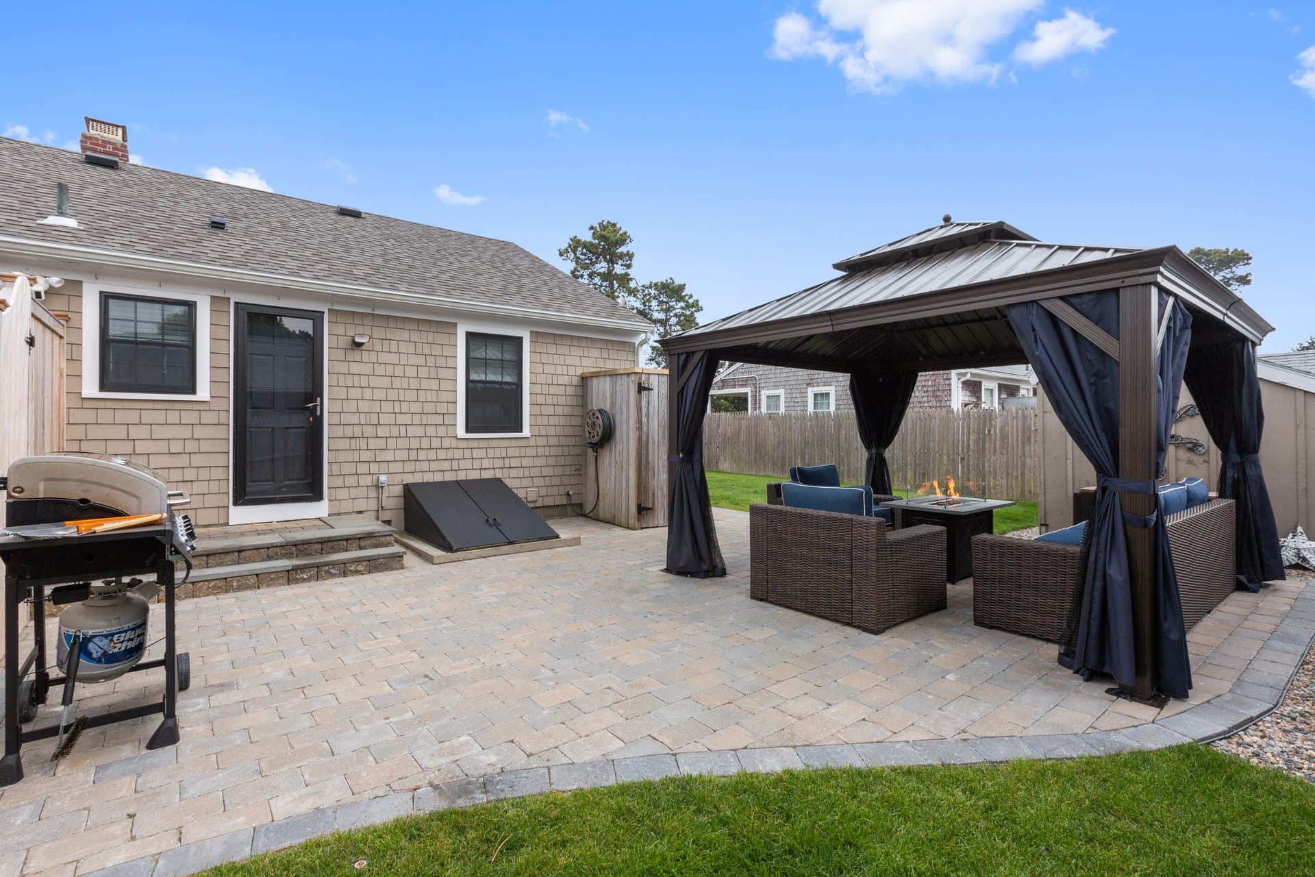 Back of a shingled house with a large paver patio, a gas grill at left and a curtained gazebo with wicker seating at right.