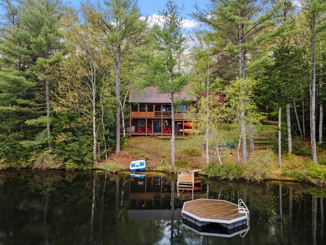 Wood cabin among tall pines seen from the lake, with a paddle boat on shore, a small dock and an octagonal swim platform.
