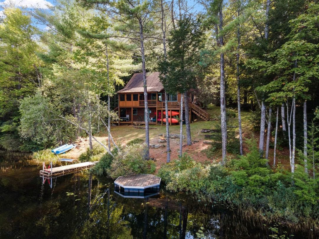 Wooden cabin with a wraparound deck among tall trees, a dock with a small boat and an octagonal floating platform on the lake.